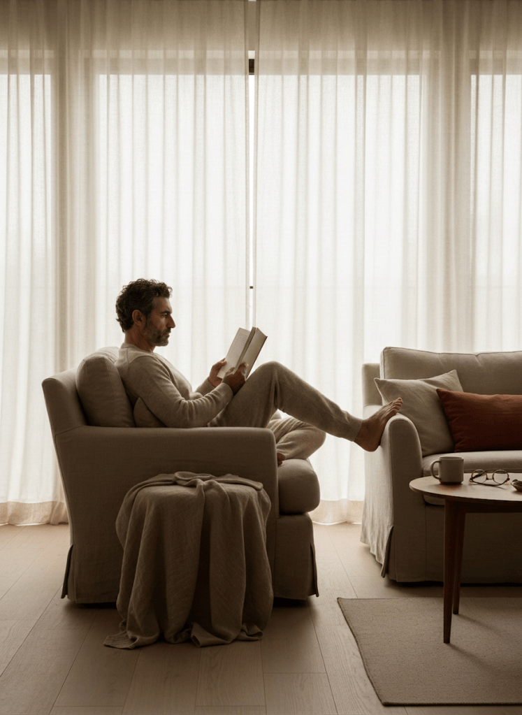 A person reading a book by a large window with soft diffused daylight, seen from the side, minimalist neutral interior, linen textures, warm muted earth tones, editorial lifestyle magazine photography, quiet contemplative mood, cream tones, no text