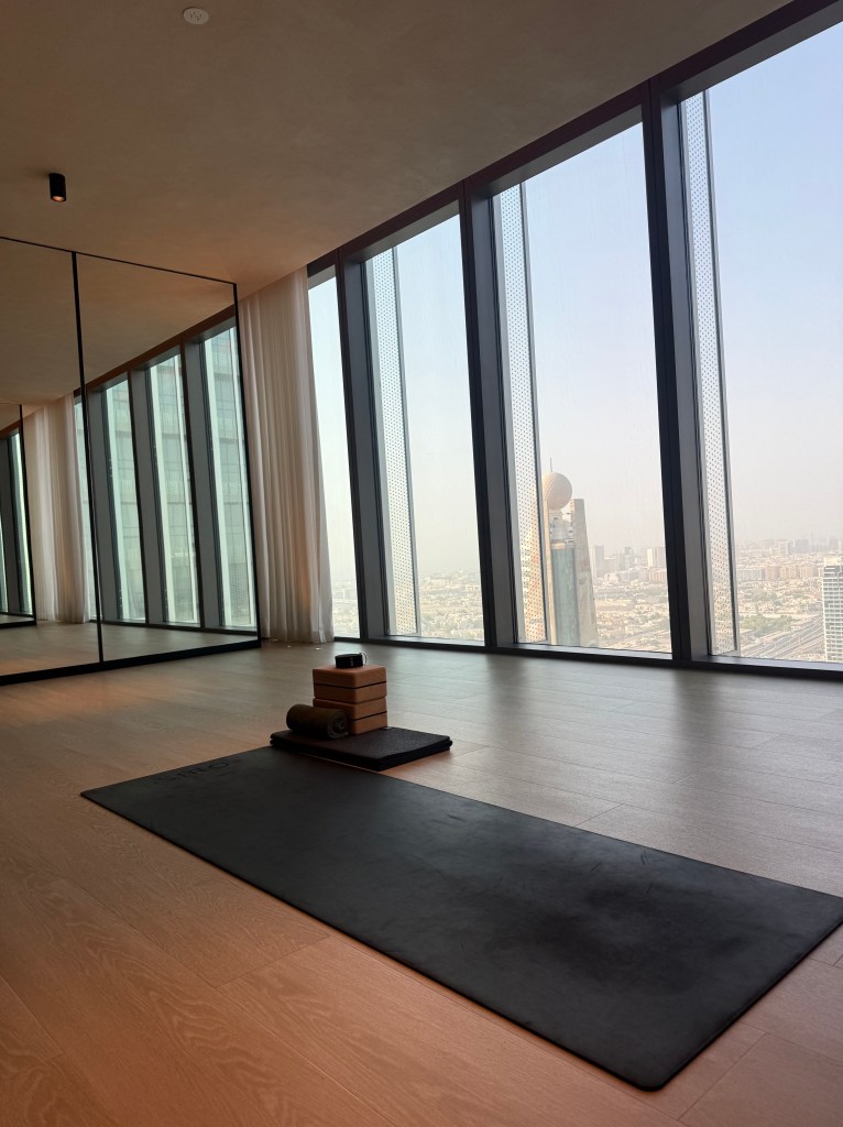 A modern yoga studio with large windows showcasing a cityscape, featuring a black yoga mat, yoga blocks, and a rolled towel on the floor.